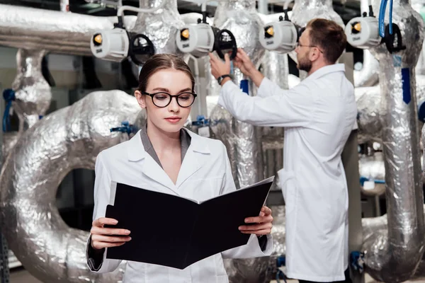 Enfoque selectivo de ingeniero atractivo en bata blanca mirando la carpeta cerca de compañero de trabajo y sistema de aire comprimido - foto de stock