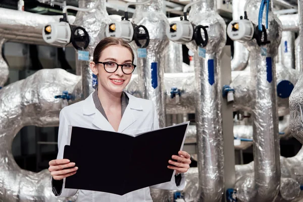 Happy and attractive engineer in white coat holding black folder near air compressed system — Stock Photo