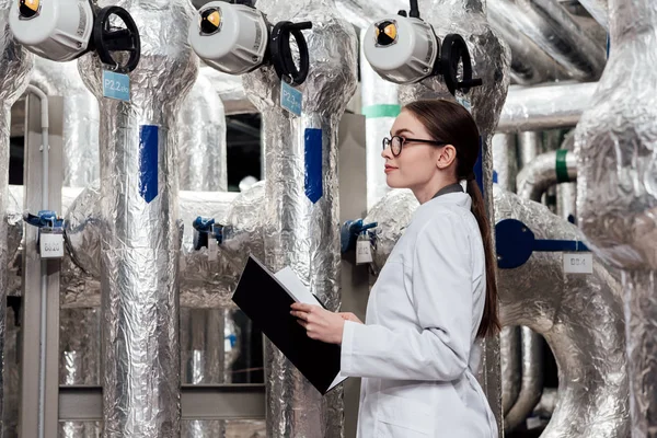 Attraktiver Ingenieur in weißem Mantel und Brille mit schwarzem Ordner in der Nähe der Druckluftanlage — Stockfoto