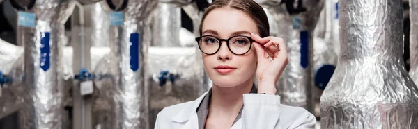 Panoramic shot of attractive engineer in white coat touching glasses near air compressed system — Stock Photo