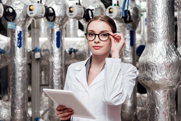 Attraktiver Ingenieur in weißem Mantel, der eine Brille berührt, während er ein digitales Tablet in der Nähe eines komprimierten Druckluftsystems hält — Stockfoto