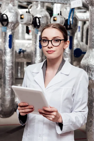 Attractive engineer in white coat and glasses holding digital tablet near air compressed system — Stock Photo