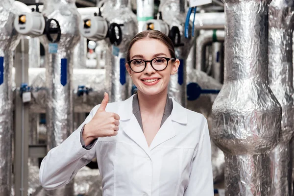 Ingeniero feliz en bata blanca y gafas que muestran el pulgar hacia arriba cerca del sistema de aire comprimido - foto de stock