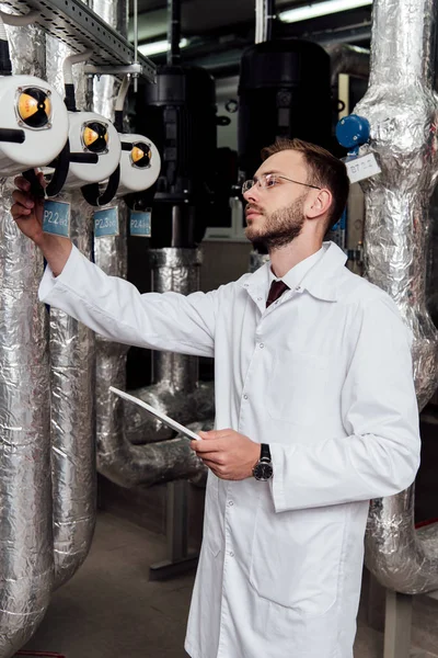 Handsome bearded engineer in white coat holding digital tablet near air supply system — Stock Photo