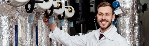 Panoramic shot of cheerful bearded engineer in white coat near air supply system — Stock Photo