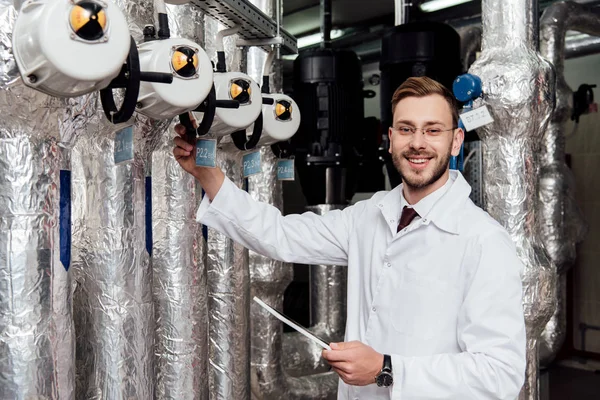 Cheerful bearded engineer in white coat holding digital tablet near air supply system — Stock Photo