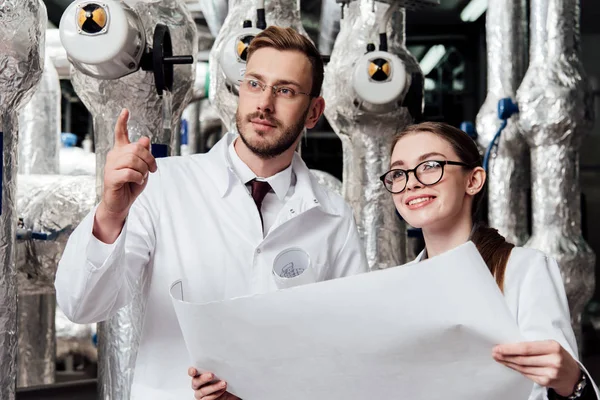 Bearded engineer in glasses pointing with finger near happy coworker holding blueprint — Stock Photo