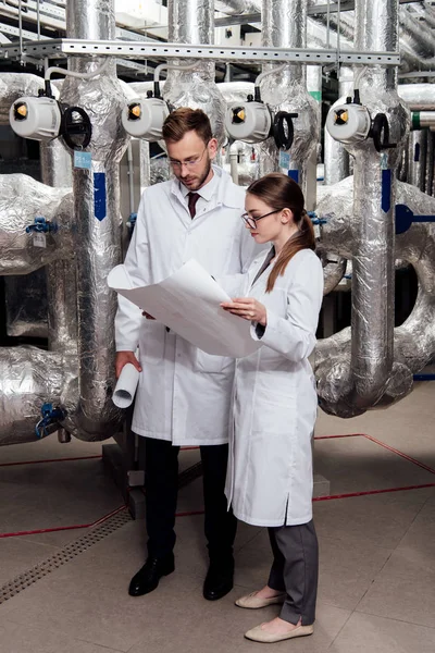 Engineers in glasses and white coats looking at blueprint near air compressor system — Stock Photo