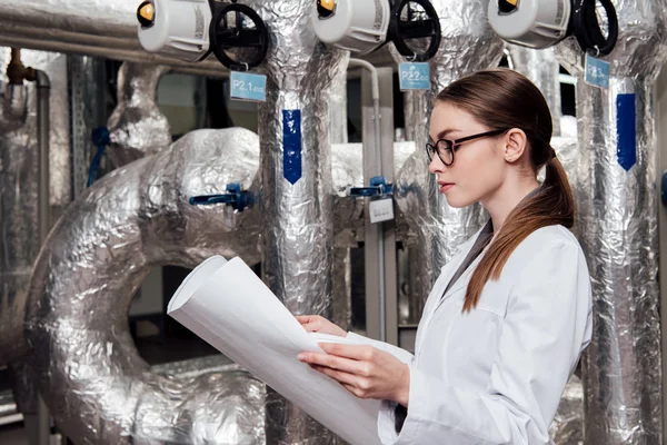 Beautiful engineer in white coat and glasses looking at blueprint near air supply system — Stock Photo