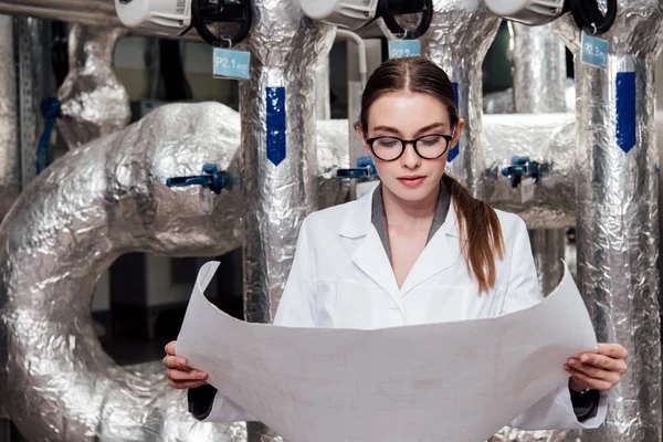 Attraktiver Ingenieur in weißem Mantel und Brille, der den Bauplan in der Nähe der Luftzufuhr betrachtet — Stockfoto