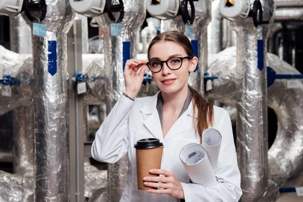 Cheerful engineer in white coat and glasses holding blueprints and paper cup near air supply system — Stock Photo