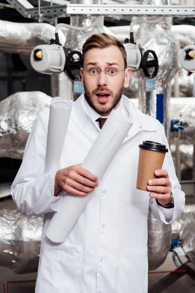 Перепуганный инженер в белом халате и очках с чертежами и бумажной чашкой возле системы подачи воздуха — стоковое фото