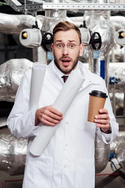 Ingeniero asustado en bata blanca y gafas que sostienen planos y taza de papel cerca del sistema de suministro de aire - foto de stock
