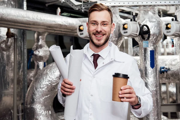 Ingeniero alegre en bata blanca sosteniendo planos y taza de papel cerca del sistema de suministro de aire - foto de stock