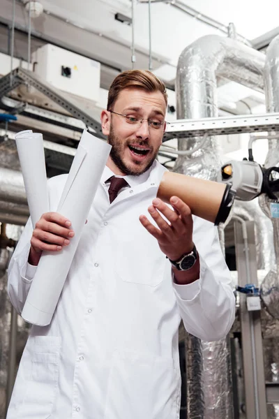 Worried engineer in white coat and glasses holding blueprints and looking at falling paper cup near air supply system — Stock Photo
