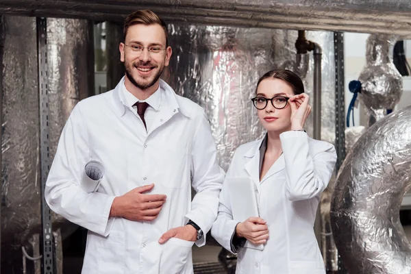 Happy scientist holding blueprints near beautiful coworker with digital tablet — Stock Photo
