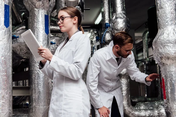 Atractivo ingeniero sosteniendo tableta digital cerca de compañero de trabajo mirando el sistema de compresor de aire - foto de stock