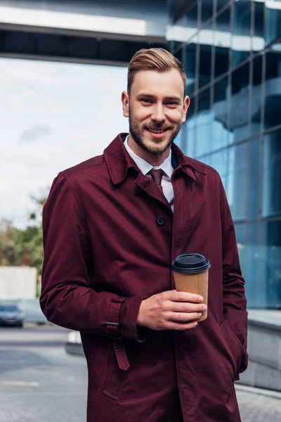 Happy bearded man in coat holding paper cup while standing with hand in pocket — Stock Photo