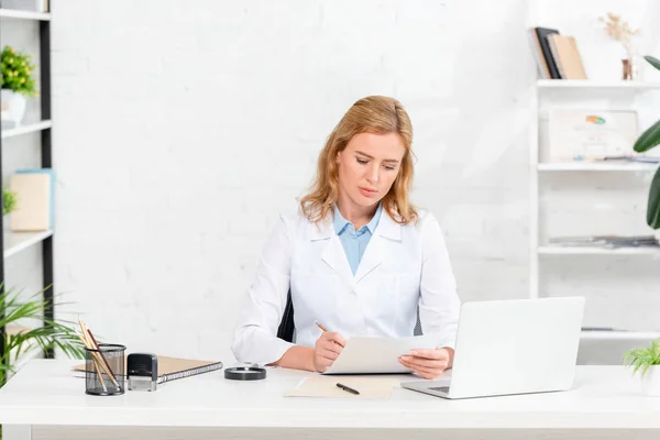 Atractiva nutricionista sentada en la mesa y escribiendo en papel en la clínica - foto de stock