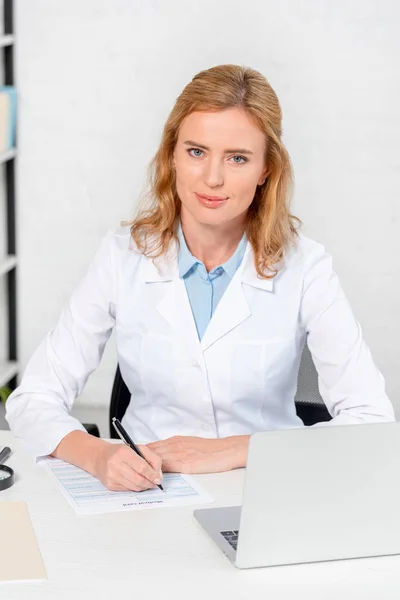 Atractiva nutricionista sentada a la mesa y sosteniendo la pluma en la clínica — Stock Photo