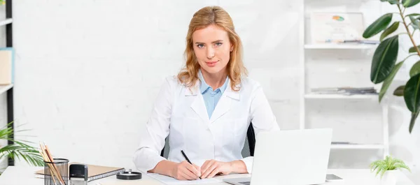 Панорамный снимок привлекательного диетолога, сидящего за столом и держащего ручку в клинике — стоковое фото