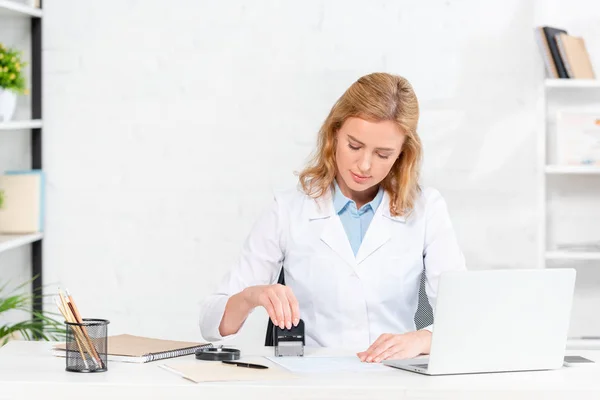 Attractive nutritionist sitting at table and stamping paper in clinic — Stock Photo