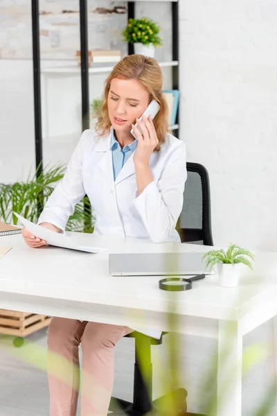 Attractive nutritionist holding paper and talking on smartphone in clinic — Stock Photo