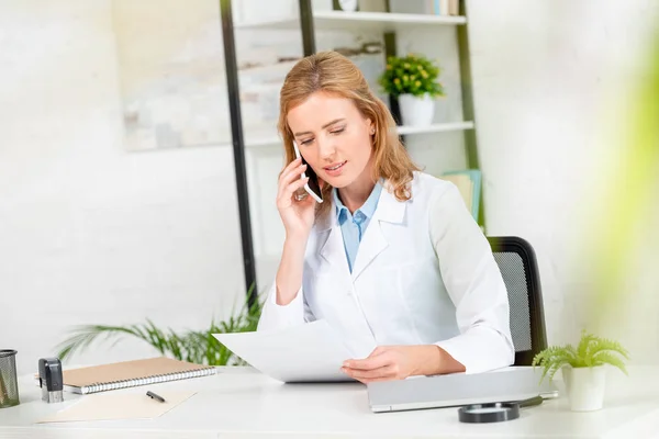 Attractive nutritionist holding paper and talking on smartphone in clinic — Stock Photo