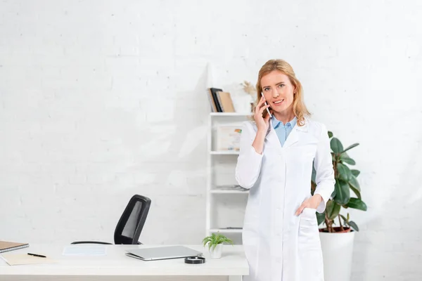 Atractiva nutricionista con la mano en el bolsillo hablando en el teléfono inteligente en la clínica - foto de stock