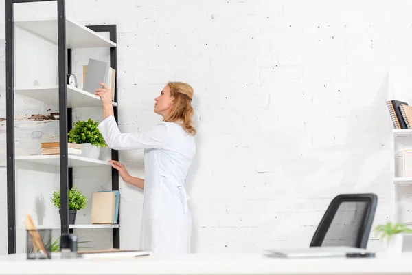 Vista laterale di attraente nutrizionista prendendo libro dalla libreria in clinica — Foto stock