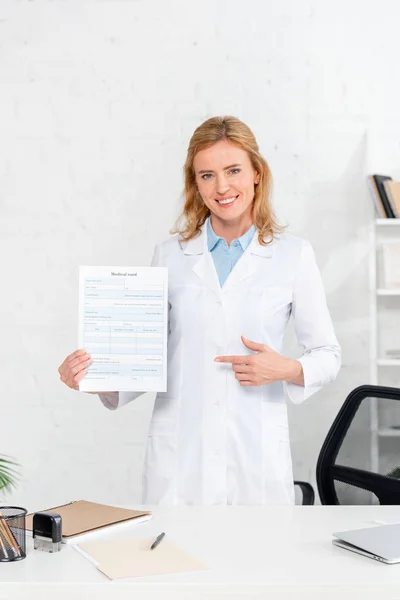 Nutricionista sonriente señalando con el dedo a la tarjeta médica en la clínica - foto de stock