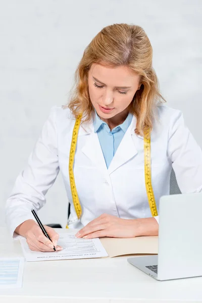 Attraktive Ernährungsberaterin sitzt am Tisch und schreibt in der Klinik auf Papier — Stockfoto