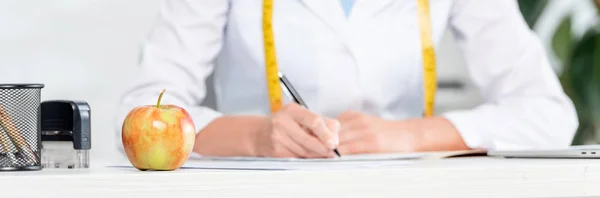 Tiro panorâmico de maçã na mesa e nutricionista sentado à mesa e escrevendo no fundo na clínica — Fotografia de Stock