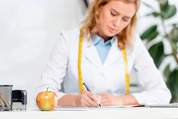 Selective focus of apple on table and nutritionist sitting at table and writing on background in clinic — Stock Photo