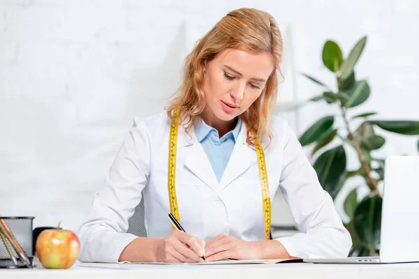 Atractiva nutricionista sentada a la mesa y escribiendo en la clínica - foto de stock