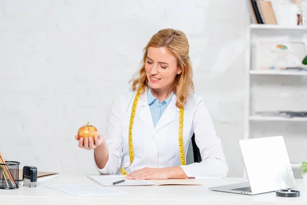 Nutritionniste souriant assis à la table et tenant pomme à la clinique — Photo de stock