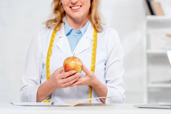 Vista ritagliata del nutrizionista sorridente seduto a tavola e che tiene la mela in clinica — Foto stock