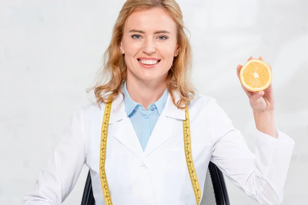 Lächelnder Ernährungsberater mit Maßband, der in der Klinik eine geschnittene Orange hält — Stockfoto