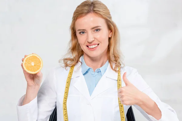 Smiling nutritionist with measure tape holding cut orange and showing like in clinic — Stock Photo