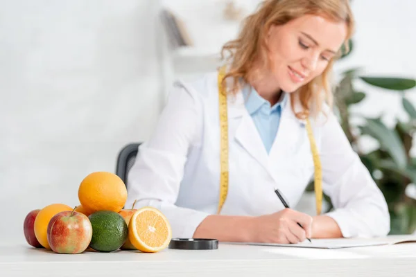 Foco seletivo de frutas e vegetais na mesa e nutricionista sentado à mesa e escrevendo em segundo plano na clínica — Fotografia de Stock