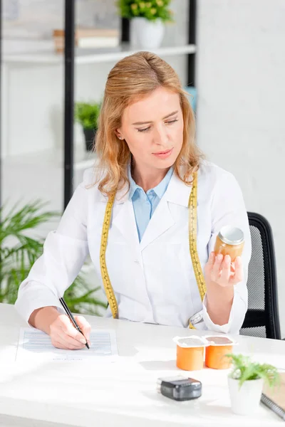 Atractiva nutricionista sentada a la mesa y sosteniendo la botella en la clínica - foto de stock