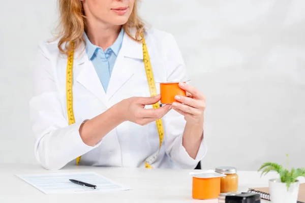 Vista cortada de nutricionista sentado à mesa e segurando iogurte na clínica — Fotografia de Stock