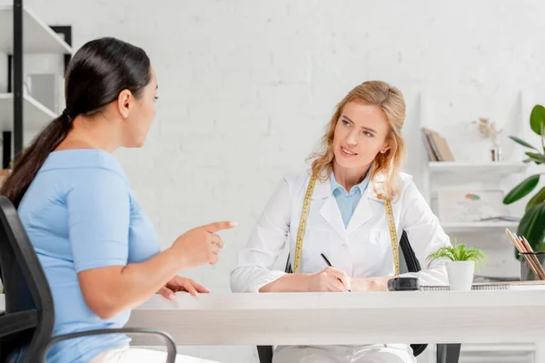 Привлекательный диетолог, сидящий за столом и разговаривающий с пациентом в клинике — стоковое фото