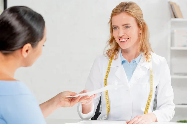 Nutricionista atractivo y sonriente sentado en la mesa y dando papel al paciente en la clínica - foto de stock