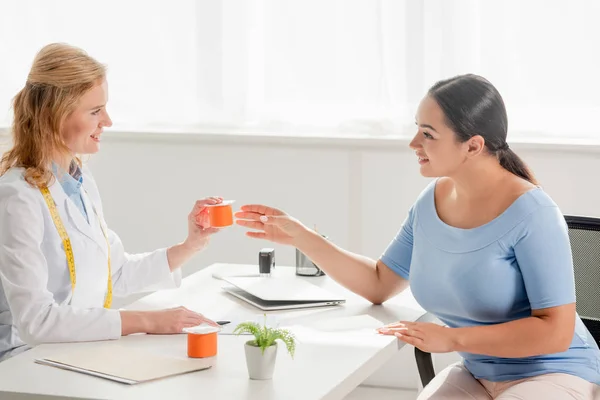 Seitenansicht des lächelnden Ernährungswissenschaftlers, der am Tisch sitzt und dem Patienten in der Klinik Joghurt reicht — Stockfoto