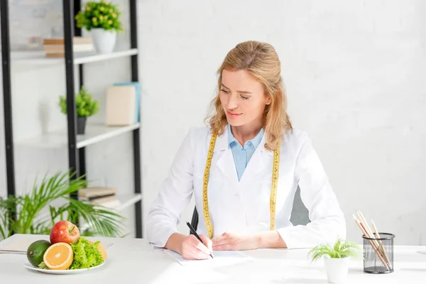 Attractive nutritionist sitting at table and looking at fruits in clinic — Stock Photo