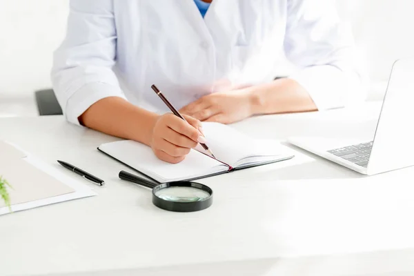 Vista recortada del dermatólogo sentado en la mesa y la escritura en el cuaderno en la clínica - foto de stock