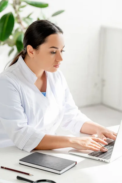 Atractivo dermatólogo sentado en la mesa y el uso de ordenador portátil en la clínica - foto de stock