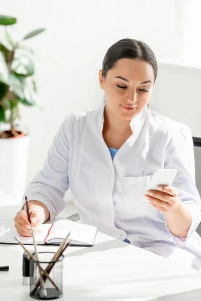 Attractive dermatologist sitting at table and using smartphone in clinic — Stock Photo