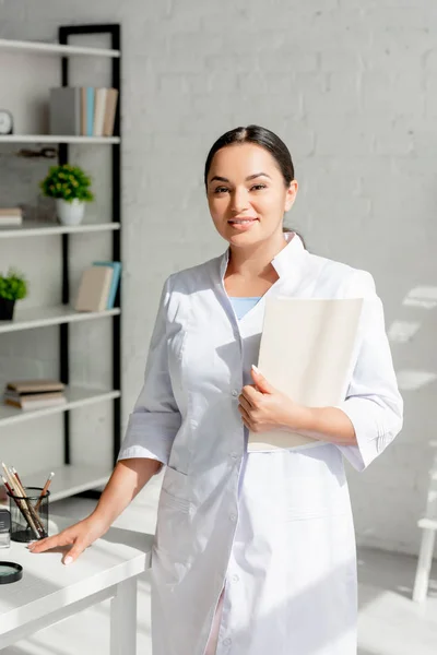 Lächelnder Hautarzt hält Mappe in der Hand und blickt in Klinik in die Kamera — Stockfoto